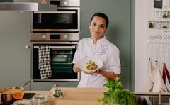 En kvinna i vit kockrock står i ett kök med gröna köksluckor. Framför henne på spisen står en kopparkastrull och glasburkar. Hon håller upp en djup vit tallrik med potatismos och pannbiffar.