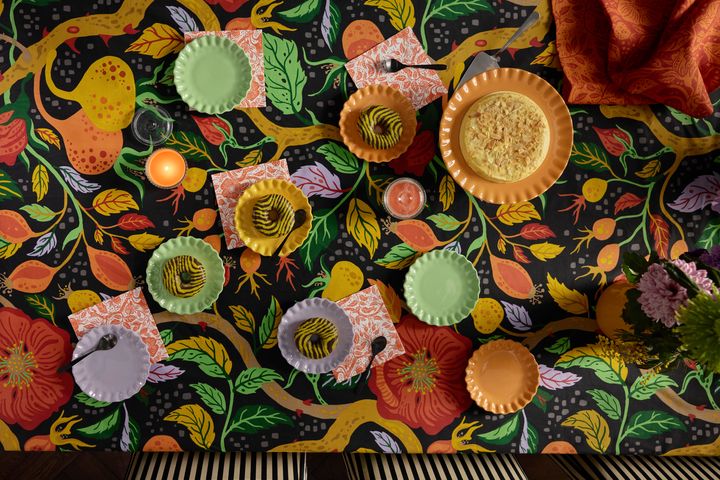En bordsduk med svart botten och mönster med färgglada blommor. På duken ligger en orange kökshadduk och det står tallrikar i grönt, lila och orange med munkar på.