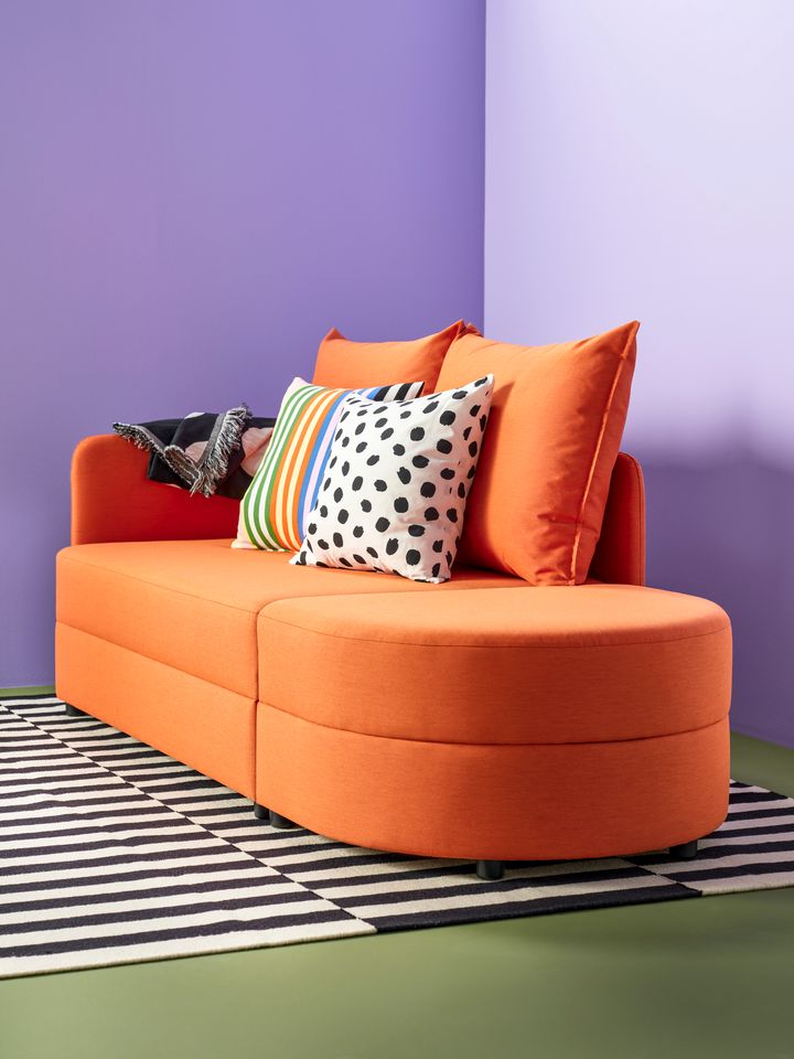 Orange%20soffa%20med%20f%E4rgglada%20kuddar%20och%20en%20svartvit%20randig%20matta%20framf%F6r%20en%20lila%20v%E4gg.