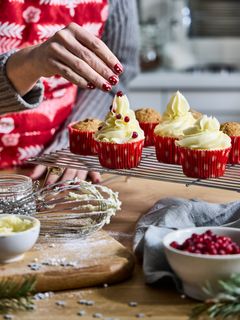 En person dekorerar julcupcakes med vita glasyrer och röda strössel.