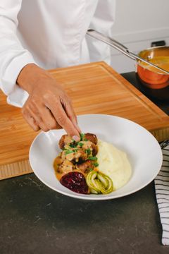 En kvinna i kockkläder strör hackad persilja över en tallrik med köttbullar och potatismos.