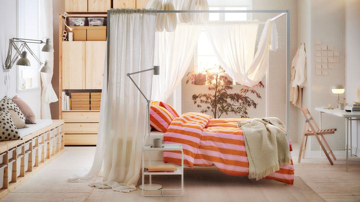 I ett sovrum står en säng med rosa och röda sängkläder. I sovrummet står trämöbler och ett vitt skrivbord.