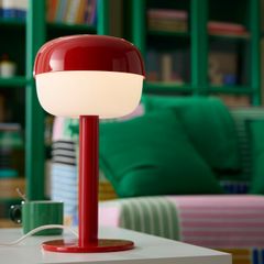 Röd bordslampa med rund skärm, tänd och placerad på ett bord framför en grön bokhylla.