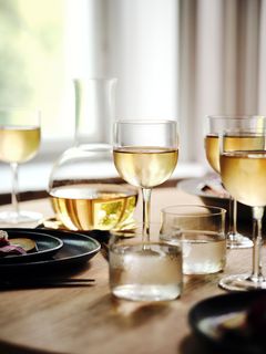 Fyra glas med vitt vin och en karaff står på ett träbord med tallrikar i bakgrunden.