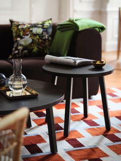 Två svarta runda bord i ett vardagsrum, med en tidning och en liten skål ovanpå. En glasvas står på ett bricka på det andra bordet. En färgglad kudde och en grön filt ligger på en brun soffa i bakgrunden.