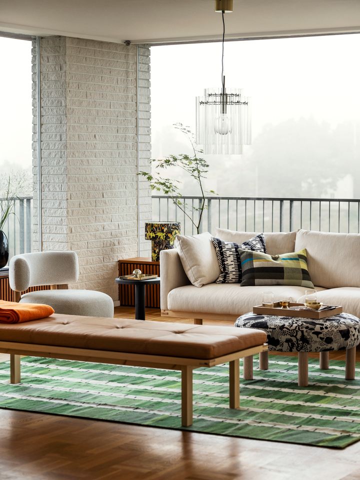 Ett%20modernt%20vardagsrum%20med%20en%20beige%20soffa%2C%20f%E5t%F6lj%2C%20tr%E4b%E4nk%20och%20ett%20rundat%20soffbord.%20Gr%F6na%20och%20bruna%20nyanser%20dominerar%2C%20med%20v%E4xter%20i%20bakgrunden%20och%20stora%20f%F6nster%20som%20sl%E4pper%20in%20naturligt%20ljus.