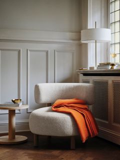 En beige fåtölj med en orange filt över ryggstödet. Bredvid finns ett runt sidobord med en guldfärgad skål. I bakgrunden står en lampa med vit skärm.