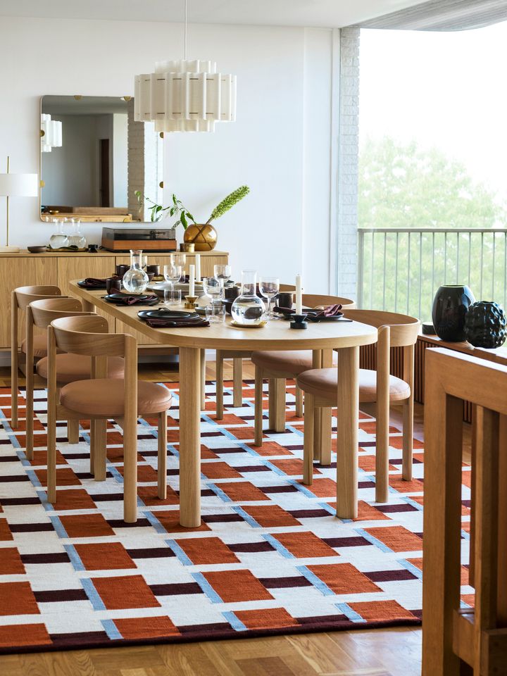 En%20modern%20matsal%20med%20ett%20l%E5ngt%20tr%E4bord%20och%20stolar.%20Bordet%20%E4r%20dukat%20med%20porslin%20och%20glas.%20Rummet%20har%20stora%20f%F6nster%20och%20ett%20geometriskt%20m%F6nstrat%20r%F6tt%20och%20vitt%20golv.