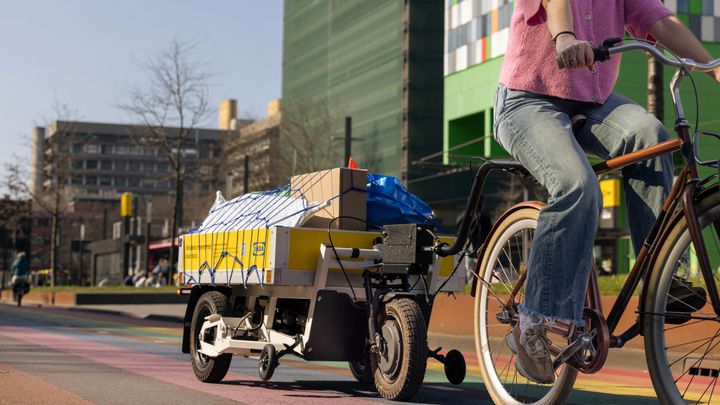 En person cyklar på en röd cykel. Efter cykeln hänger ett vitt släp lastat med IKEA-produkter.