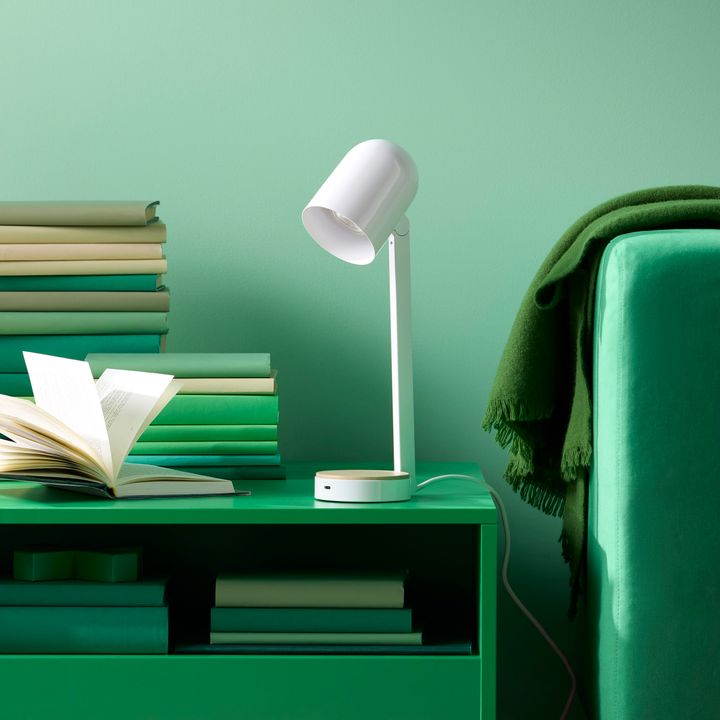 En%20vit%20skrivbordslampa%20p%E5%20ett%20gr%F6nt%20bord%20med%20en%20h%F6g%20b%F6cker%20och%20en%20gr%F6n%20f%E5t%F6lj%20i%20bakgrunden.