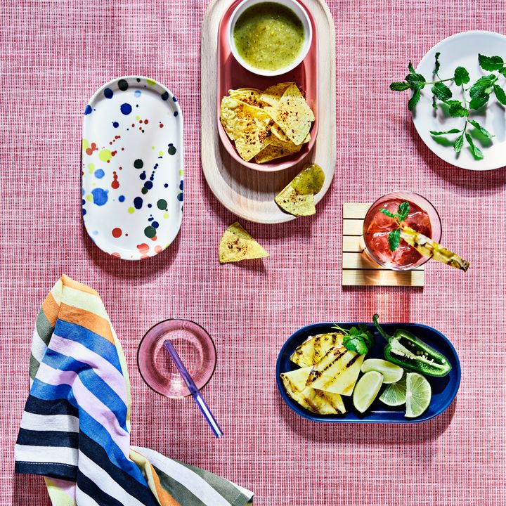 En%20samling%20f%E4rgglada%20fat%20med%20chips%2C%20s%E5s%2C%20grillad%20frukt%2C%20en%20drink%20och%20en%20randig%20servett%20p%E5%20ett%20rosa%20underlag.