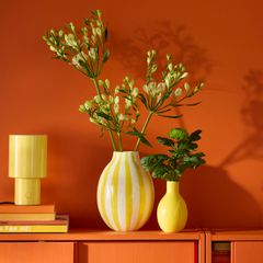 Två vaser, en randig och en enfärgad gul, med blommor på en orange hylla mot en orange vägg. En gul lampa och böcker finns bredvid.
