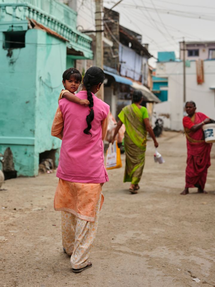 En%20kvinna%20i%20rosa%20kl%E4der%20b%E4r%20ett%20barn%20och%20g%E5r%20p%E5%20en%20gata%20med%20fler%20m%E4nniskor%20i%20f%E4rgglada%20kl%E4der.