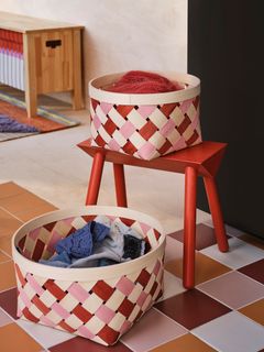 Två flätade korgar i röd och rosa nyans, en på en röd bänk och en på golvet, fyllda med textil.