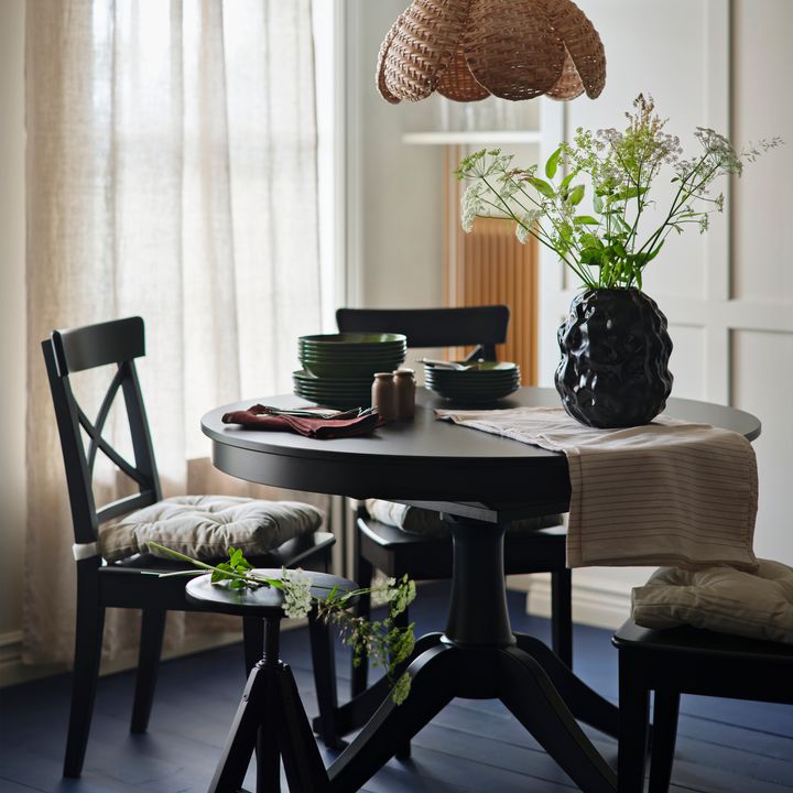 Matbord%20med%20svarta%20stolar%2C%20dukat%20med%20gr%F6na%20tallrikar%2C%20en%20vas%20med%20blommor%20och%20en%20fl%E4tad%20taklampa%20ovanf%F6r.