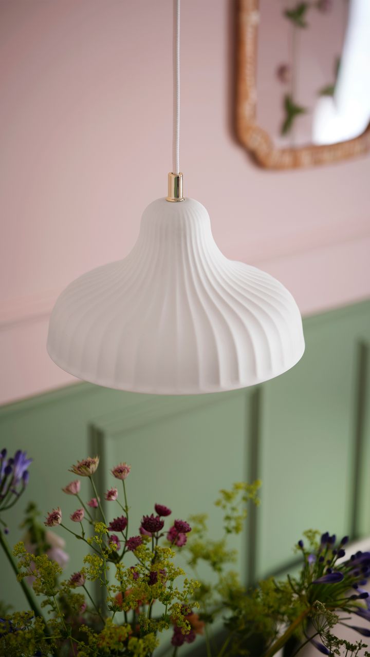 En%20vit%2C%20veckad%20taklampa%20h%E4nger%20%F6ver%20f%E4rgglada%20blommor%20i%20ett%20rum%20med%20gr%F6na%20panelv%E4ggar.