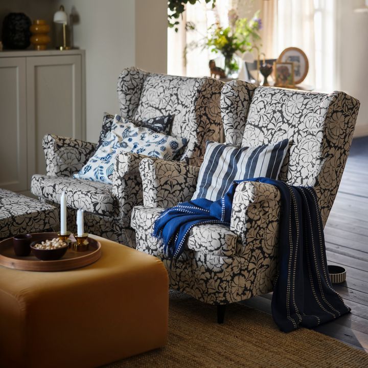 Tv%E5%20blommiga%20f%E5t%F6ljer%20med%20randiga%20och%20bl%E5%20kuddar%2C%20en%20bl%E5%20filt%2C%20samt%20ett%20brunt%20soffbord%20med%20ljus%20och%20sk%E5lar.