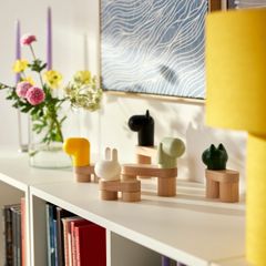 En vit hylla med färgglada, abstrakta djurfigurer och blommor i vas. Detaljer av en konsttavla syns i bakgrunden.