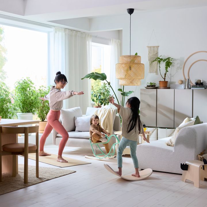 Tre%20barn%20leker%20i%20ett%20ljust%20vardagsrum%20med%20v%E4xter%20och%20modern%20inredning.%20Ett%20barn%20sitter%2C%20de%20andra%20tv%E5%20g%F6r%20r%F6relser%20p%E5%20en%20balansbr%E4da%20och%20yogamatta.