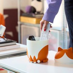 En person lägger en fjärrkontroll i en vit och orange behållare formad som en katt på ett bord.