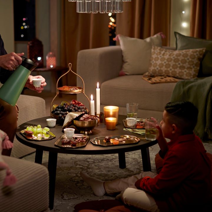 En%20mysig%20kv%E4llsscen%20med%20tv%E5%20personer%20som%20dricker%20te%20vid%20ett%20soffbord%20fyllt%20med%20snacks%20och%20frukt%2C%20omgivna%20av%20levande%20ljus.