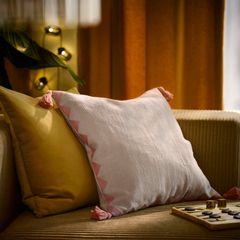 En vit kudde med rosa tofsar vilar mot en gul kudde på en beige soffa. En växt och ett brädspel syns i bakgrunden.