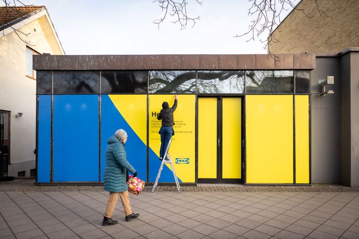 En person på stege som målar ett fönster på en byggnad i gula och blå färger. En annan person går förbi på trottoaren.