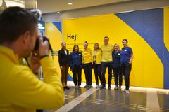 Grupp av personer i gula och blå kläder står framför en gul vägg med texten "Hej!". En person fotograferar dem.