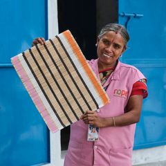 Kvinna i rosa uniform håller upp en vävd matta framför en blå dörr.