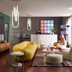 Ett modernt hotellrum med en gul soffa, vita fåtöljer, och ett runt bord. Ljusa lampor hänger från taket och en väggdekoration med färgglada cirklar syns i bakgrunden.