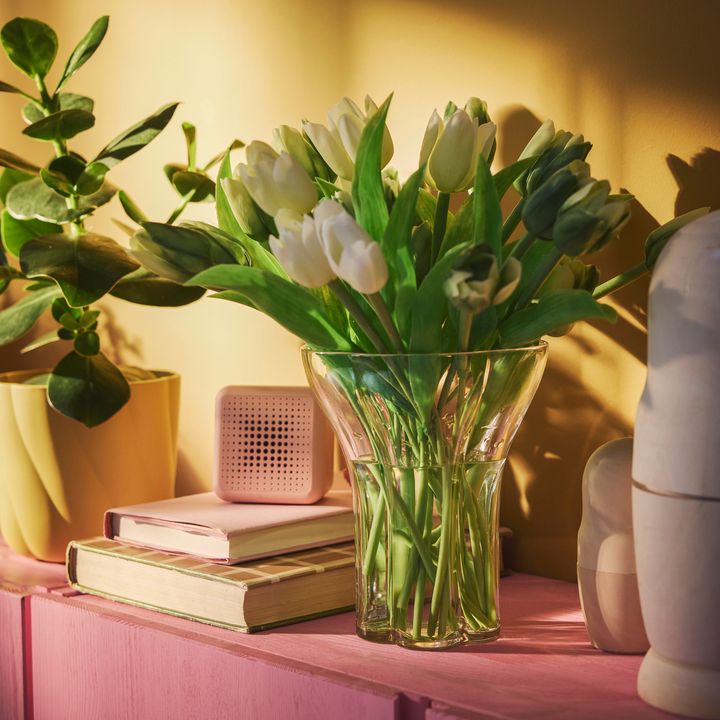 En%20bukett%20vita%20tulpaner%20i%20en%20glasvas%20p%E5%20en%20rosa%20yta%2C%20bredvid%20n%E5gra%20b%F6cker%20och%20en%20liten%20h%F6gtalare.
