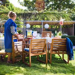 Kvinna vid dukat trädgårdsbord med trämöbler, grönska i bakgrunden.