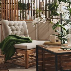 En bekväm stol med ljus dyna och en grön filt står på en träbalkong omgiven av växter. Ett bord med kaffekopp och vattenkanna framför.