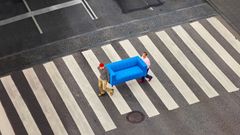 Två personer går på ett övergångsställe. De bär på en blå soffa.