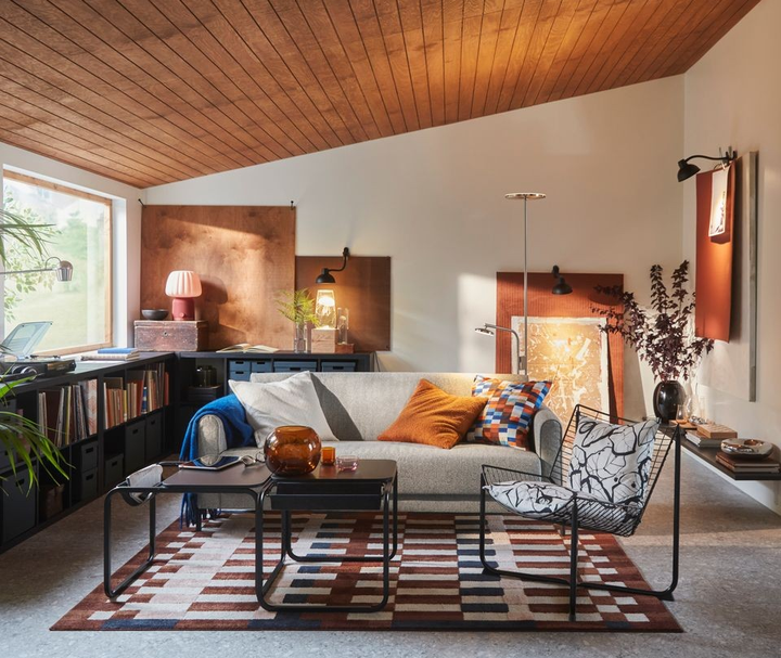 Ett modernt vardagsrum med en grå soffa, ett mönstrat soffbord, och en dekorativ matta. Det finns växter i krukor och tavlor lutade mot väggen.