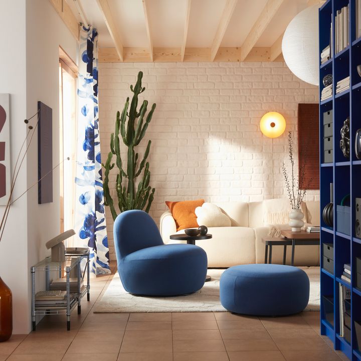 Modernt%20vardagsrum%20med%20vit%20tegelv%E4gg%2C%20beige%20soffa%2C%20bl%E5%20f%E5t%F6lj%20och%20fotpall%2C%20samt%20en%20stor%20kaktus%20i%20h%F6rnet.