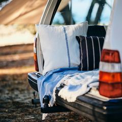 Bakluckan på en vit bil är öppen och inredd med kuddar och en filt, parkerad i ett soligt skogsområde.