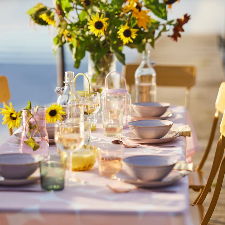 Ett%20dukat%20bord%20med%20sommarblommor%20och%20solrosor%20i%20en%20vas%2C%20omgiven%20av%20tallrikar%2C%20glas%20och%20karaffer%20under%20bl%E5%20himmel.