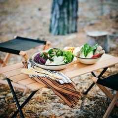 Ett träbord utomhus med skålar fyllda med sallad och färska grönsaker, omgiven av fällstolar.