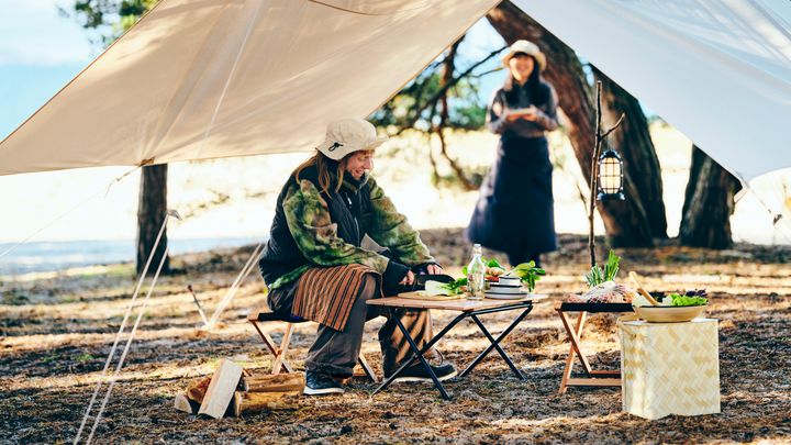 En person sitter under ett tält vid ett campingbord med mat. En annan person står i bakgrunden.