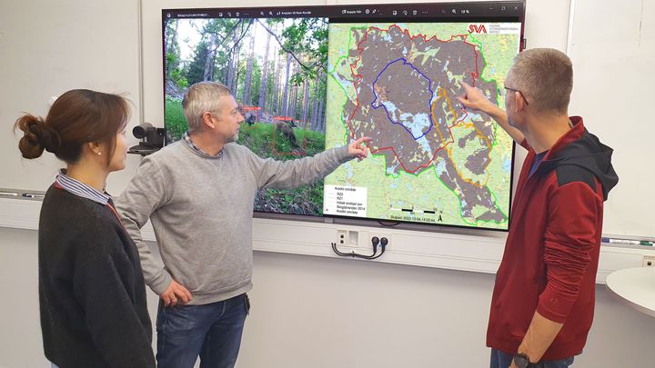 Två män och en kvinna diskuterar en karta på en stor tv-skärm.