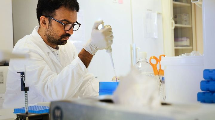 Forskare i labbrock sitter vid labbbänk med pipettverktyg i handen.