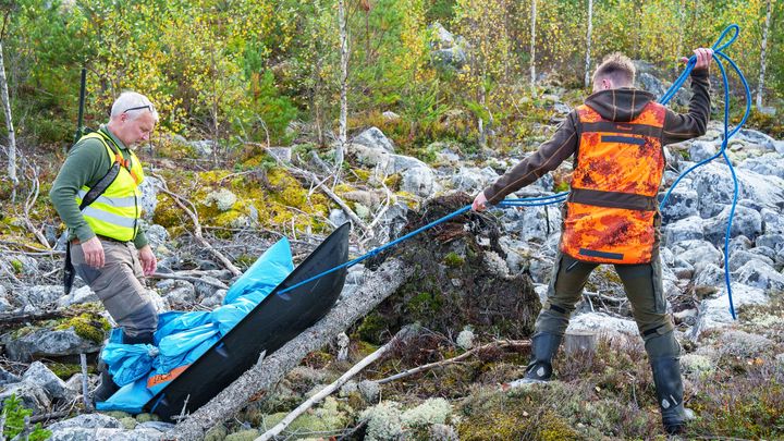 Två jägare transporterar något i en blå säck i en pulka över stock och sten.