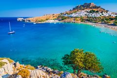 Bild på en pittoresk strand och kustlinje i Rhodos, Grekland, med turkost hav och en bergsby i bakgrunden.