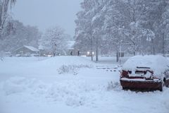 En bil täckt av snö parkerad vid ett snöigt vinterlandskap med träd och hus i bakgrunden.