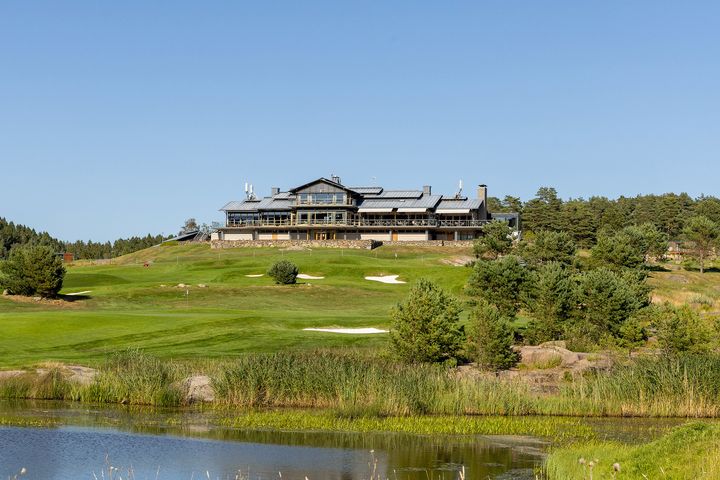 En stor byggnad på en kulle omgiven av en grön golfbana och träd, med en klarblå himmel i bakgrunden.