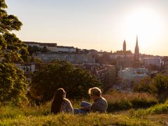 Två personer sitter i gräset och njuter av solnedgången med en utsikt över staden.