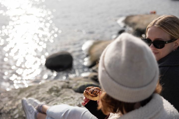 Två personer sitter på klippor vid vattnet. En av dem håller en kanelbulle.