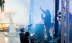 En person sjunger på en scen vid vattnet, med en segelbåt i bakgrunden.