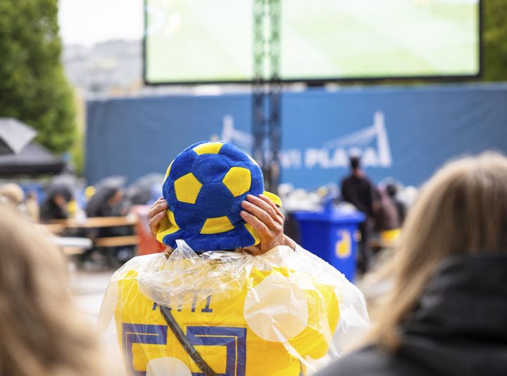 Person med gul och blå fotbollskeps ser på en stor utomhusskärm under ett evenemang.